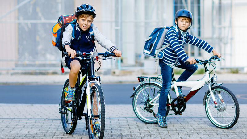 Zwei Schulkinder mit Fahrradhelm und Rucksack sitzen auf ihren Fahrrädern und blicken in die Kamera.