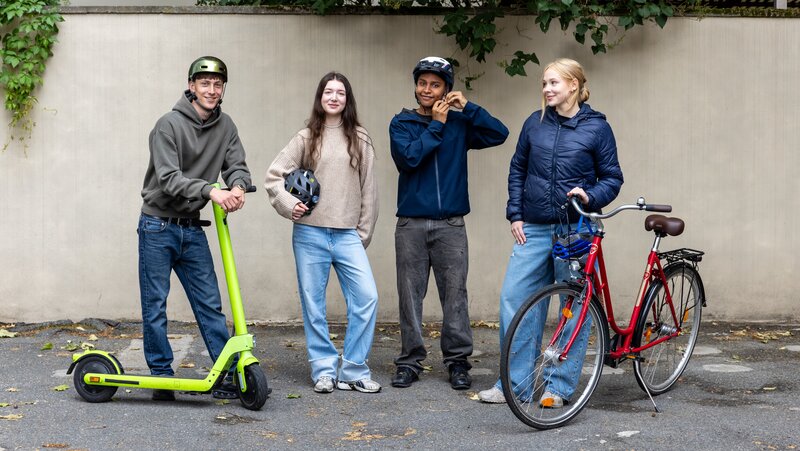 Vier junge Menschen stehen nebeneinander. Einer setzt sich gerade den Helm auf. Ein anderer hält einen E-Scooter fest.