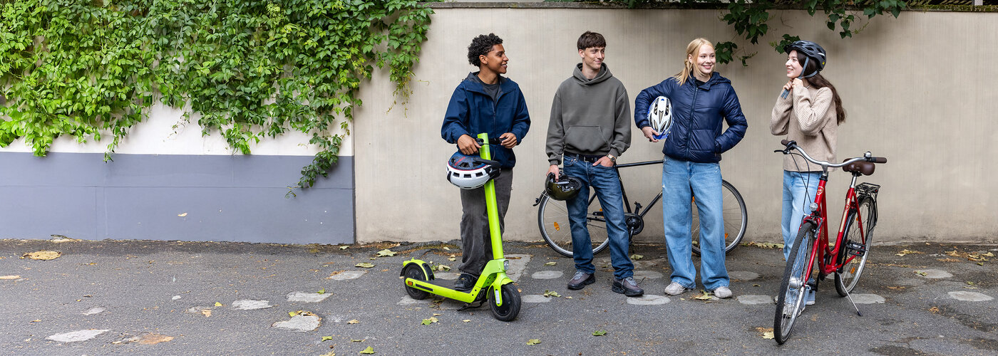 Vier Jugendliche stehen mit Fahrrad, E-Scooter und Helmen in der Hand vor einer Mauer.
